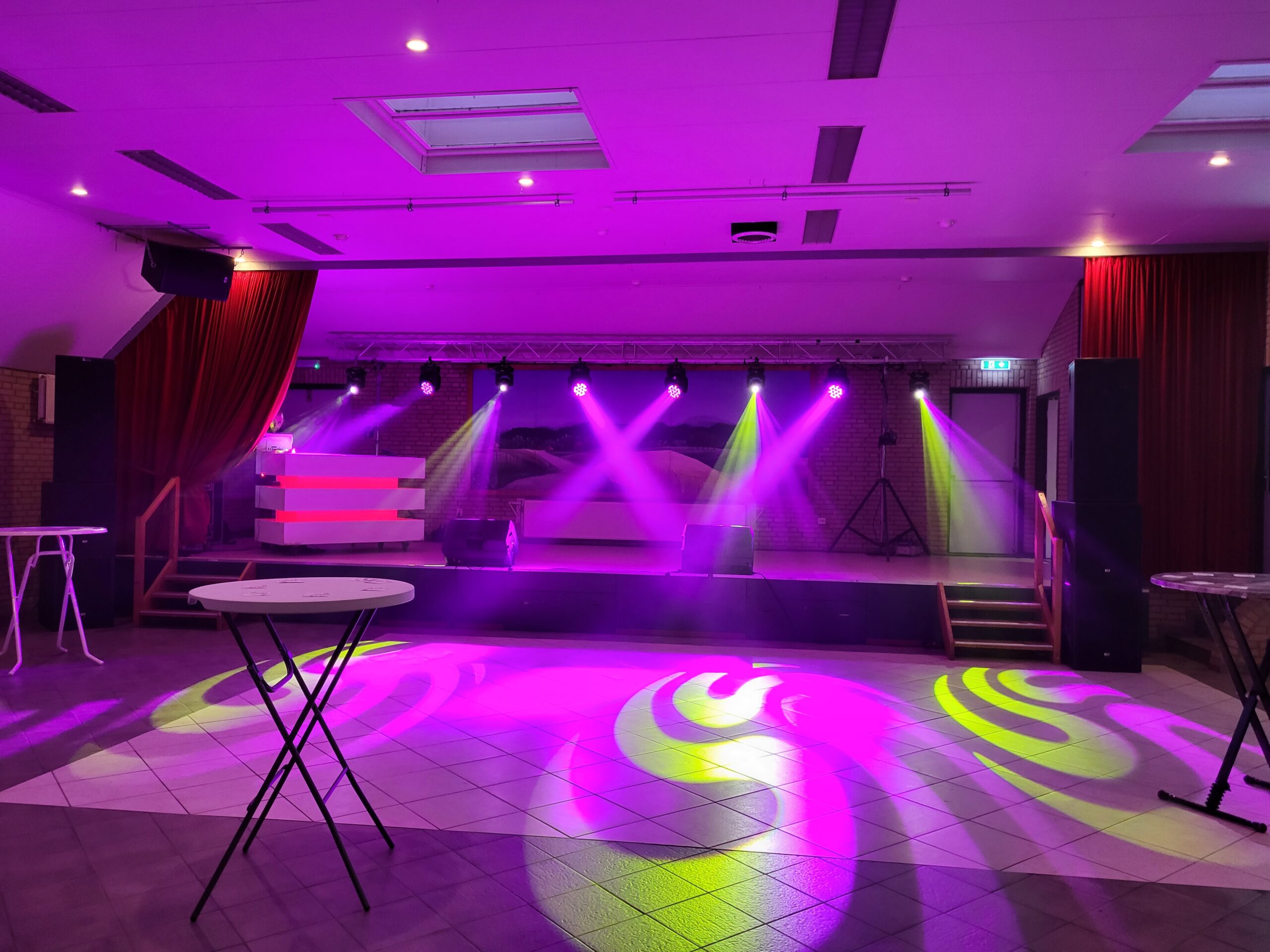 Een foto van het podium van de Foute Party 2023 in een lege zaal. Met roze en groene lichtstralen.